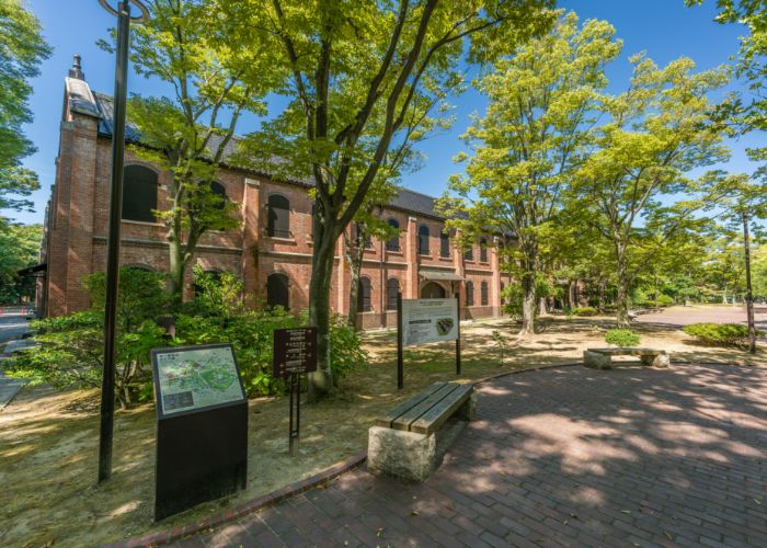 A path outside of the Ishikawa Prefectural Museum of Art, lined with green trees, benches, and a map. In the background, the red-brick museum stands tall.